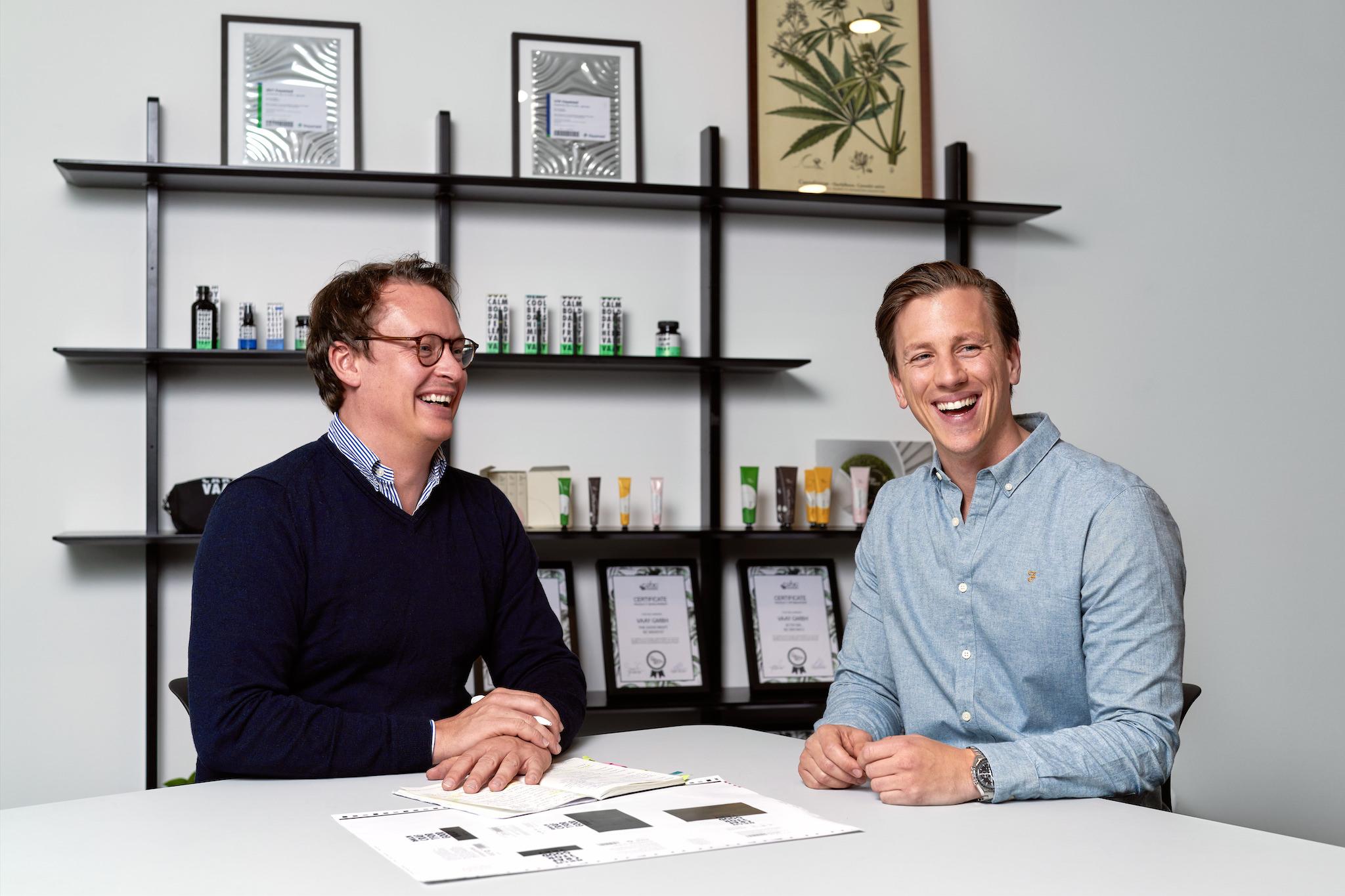 The picture shows two white men sat at a white table, both of whom are laughing. They are the founders of medical cannabis company, Sanity Group. In the background you can see pictures of cannabis plants.