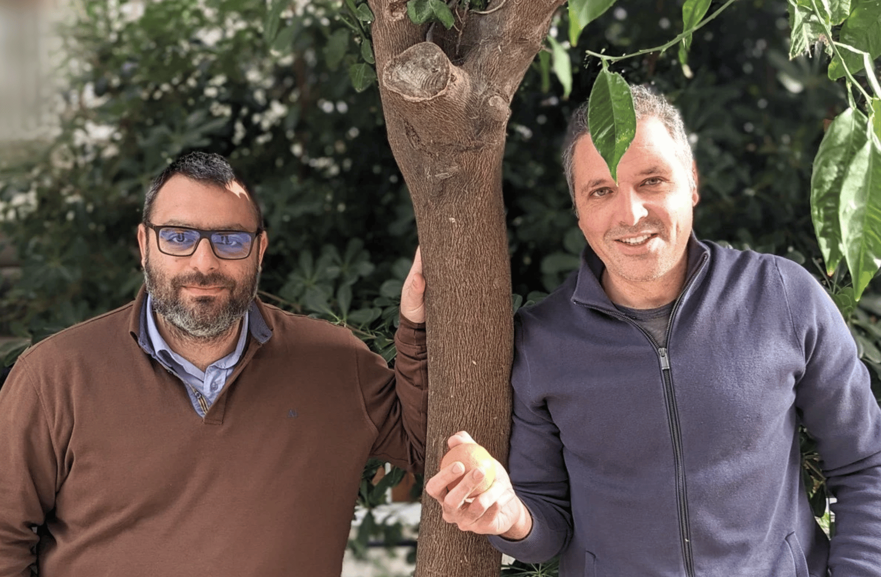 Two men standing beside a tree. One holds a piece of fruit in his hand.