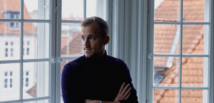 Lars Maaloe, cofounder of Corti, standing near a window with houses visible in the background.