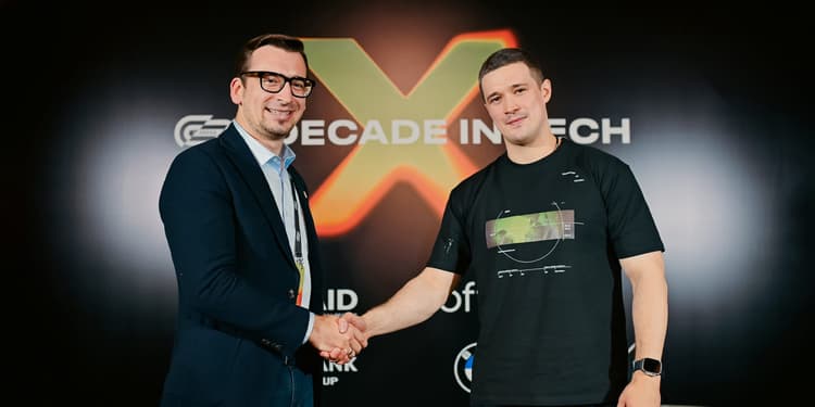 Two men shaking hands and looking at the camera. Behind them is a black background with an orange X.