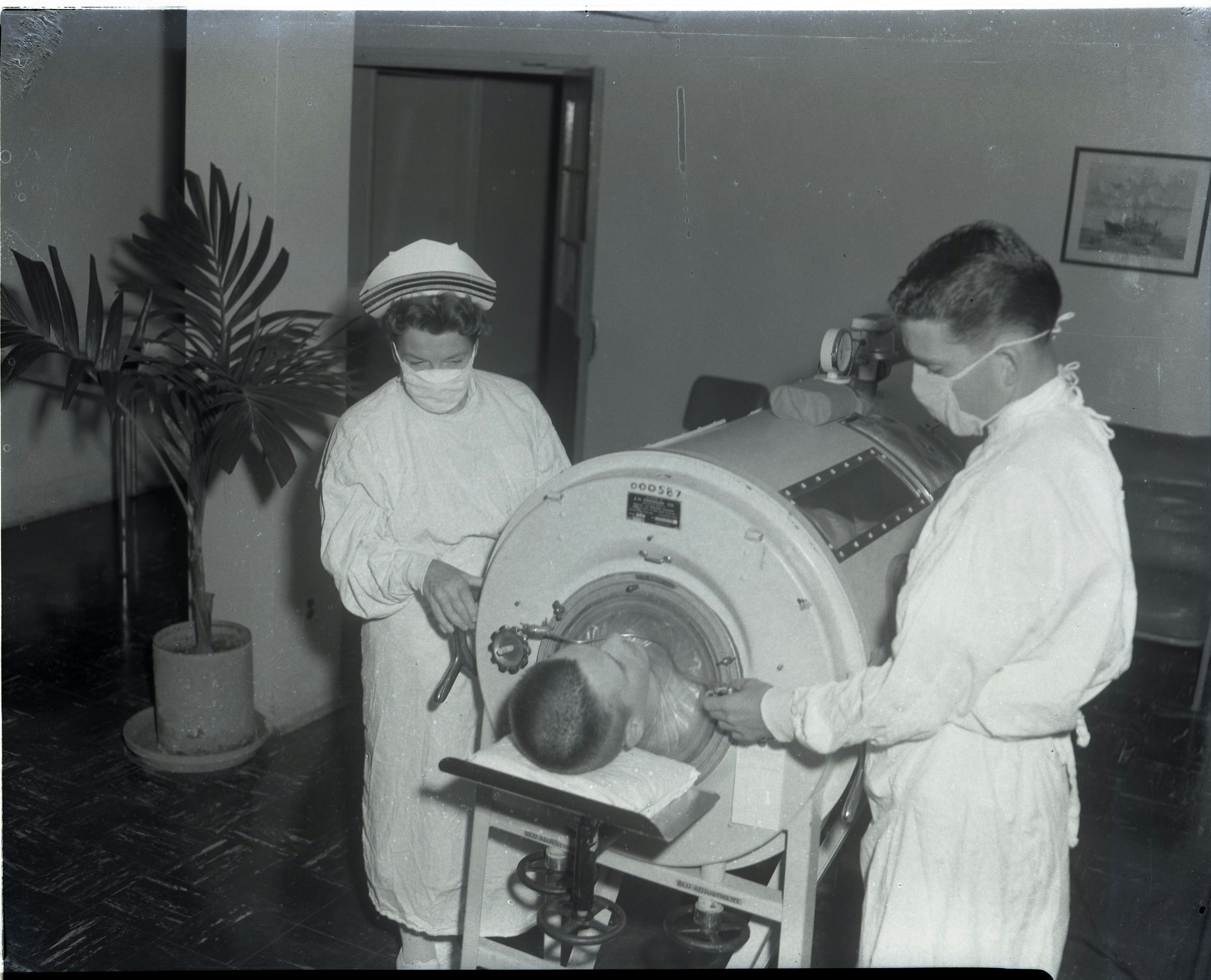 Image of a boy in an Iron Lung at the Naval Hosptial C 1960, and a doctor and a nurse standing around it. The Iron Lung was a healthcare innovation that saved many lives.