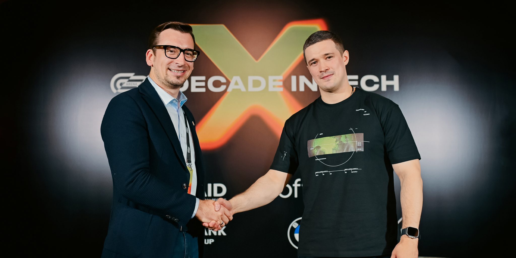 Two men shaking hands and looking at the camera. Behind them is a black background with an orange X.