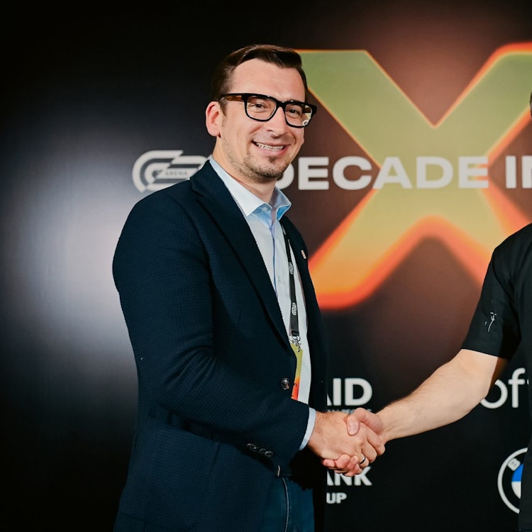 Two men shaking hands and looking at the camera. Behind them is a black background with an orange X.