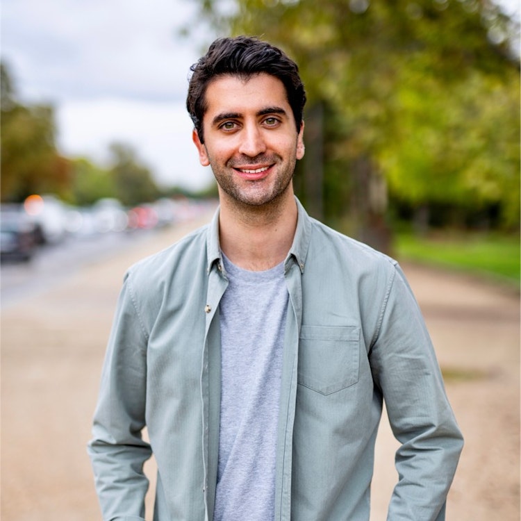 A man wearing a grey tshirt and green jacket stands in a street.