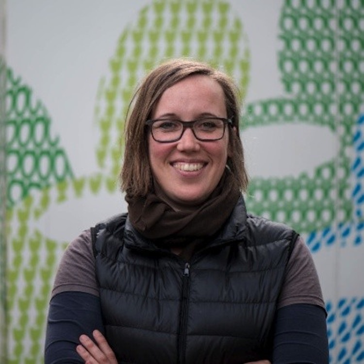 A woman wearing glasses smiles in front of a green and white wall.