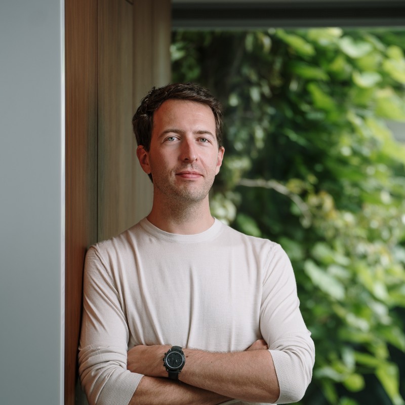 A man standing with his arms folded. Behind him is a window into some greenery.
