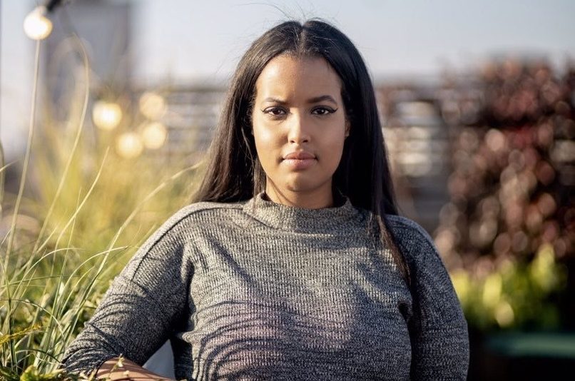 A lady with long brown hair wearing a grey jumper looks directly into the camera with a neutral look on her face.