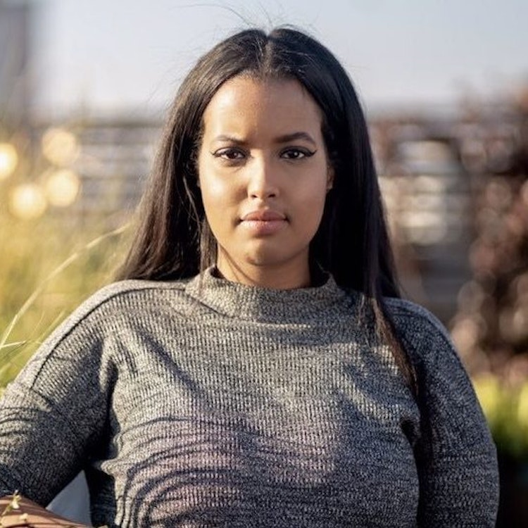 A lady with long brown hair wearing a grey jumper looks directly into the camera with a neutral look on her face.